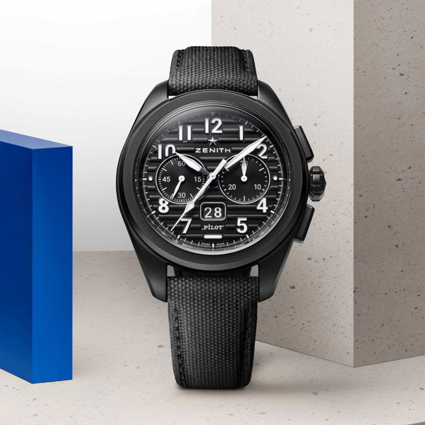 Black Zenith watch with chronograph features, displayed near blue and light gray geometric shapes.